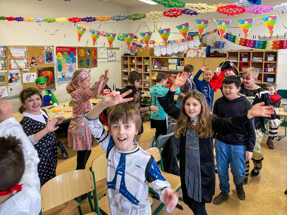 Kinder mit Kostümen feiern Fasching im Klassenraum, tanzen um Stühle, bunte Girlanden und Wimpel sind geschmückt